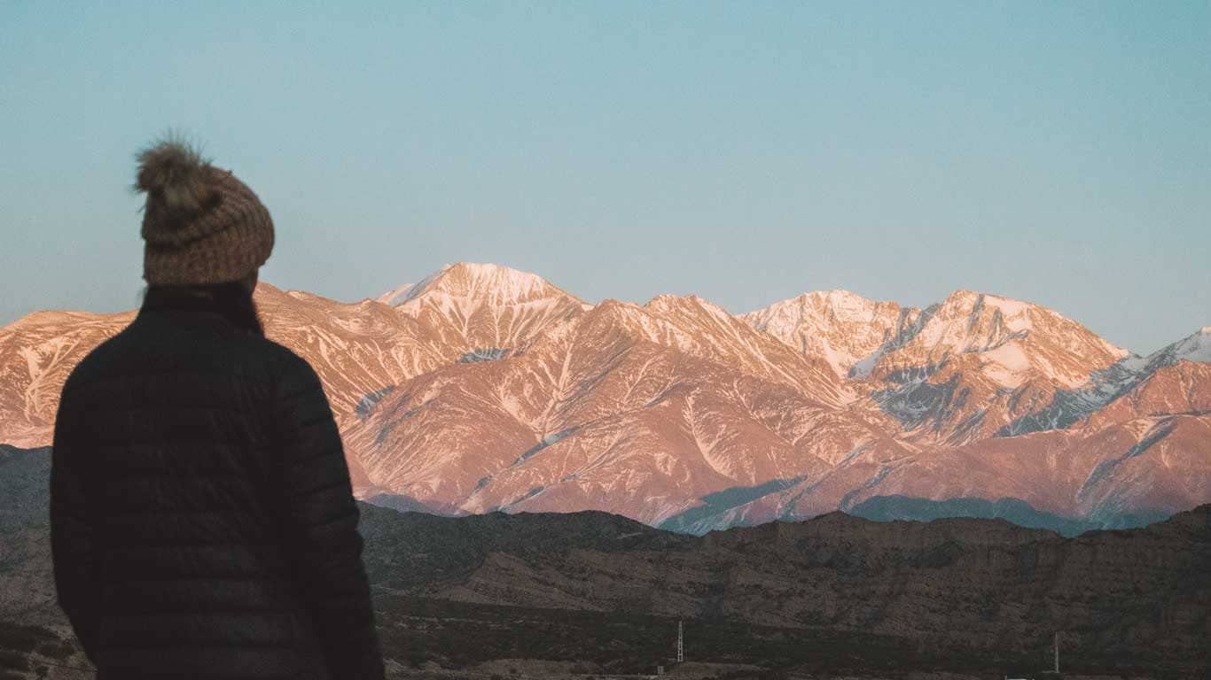 A solitary figure in winter attire stands in contemplation, gazing at the sun-kissed peaks of the Andes in Mendoza, with the orange hues of dawn or dusk painting the snow-capped mountains.