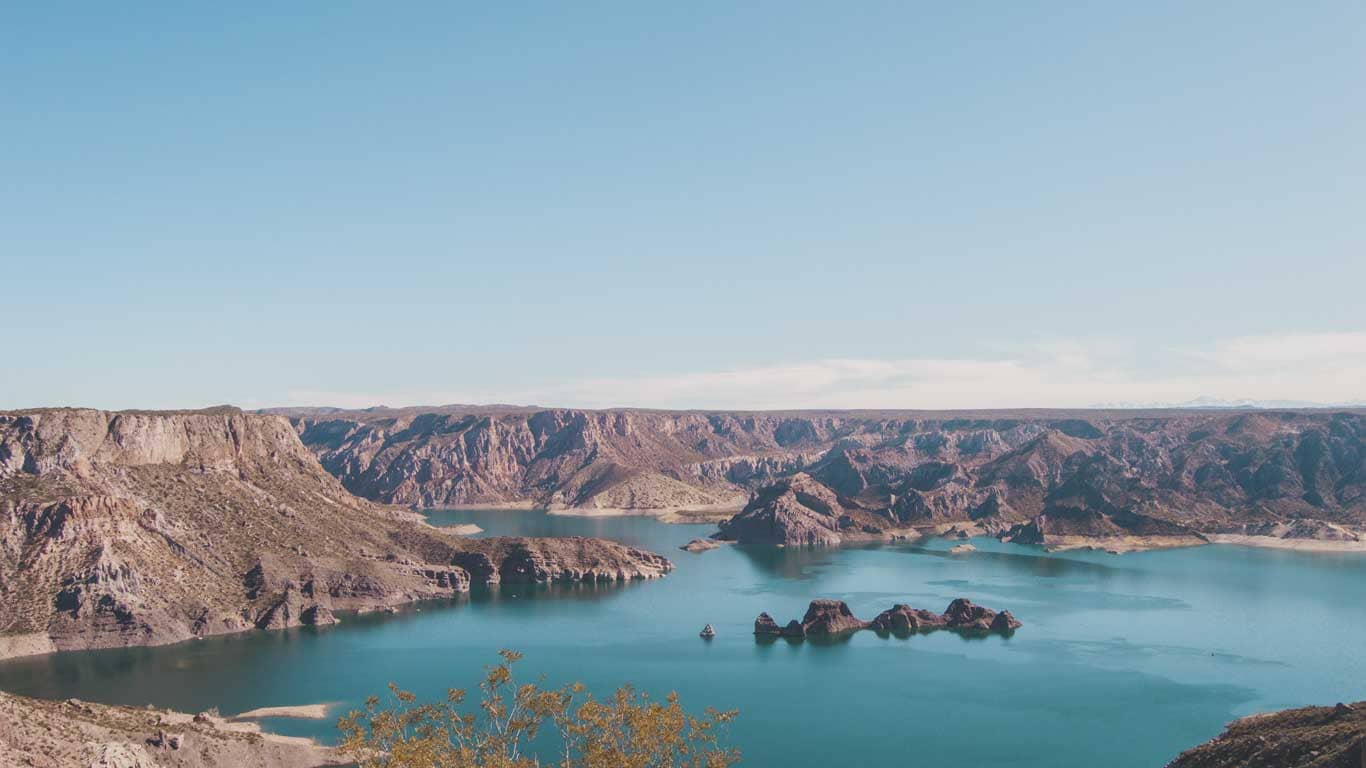 A panoramic view of Atuel Canyon in Mendoza in February day, showcasing the serene turquoise waters nestled between rugged brown cliffs under a vast blue sky.