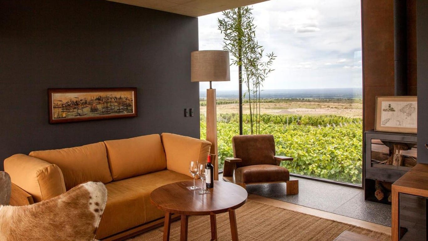 Inside the Casa de Uco hotel in Mendoza, an elegant living space with a plush mustard sofa and a brown leather armchair invites relaxation, with a view of the lush vineyards outside the large window, accompanied by a bottle of wine and two glasses ready to enjoy.