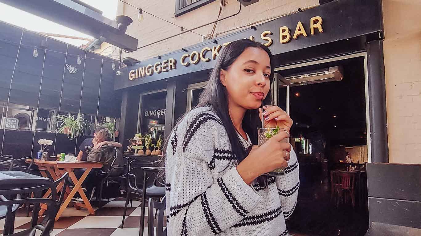 A woman enjoying a drink at Ginger's Bar in Mendoza, with the bar's name visible in the background. She is casually dressed in a patterned sweater, and the bar's outdoor seating area has a relaxed atmosphere with other patrons and plants in the background.