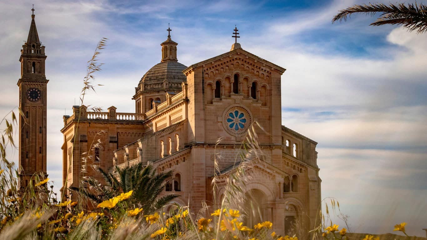 The Ta' Pinu Basilica in Gozo, Malta, stands majestically amid the wildflowers, with its ornate architecture and twin bell towers reaching towards a soft blue sky, exuding a sense of peace and spiritual grandeur.