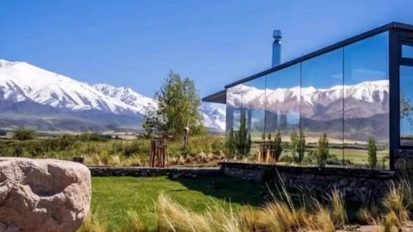 A modern hotel with expansive glass walls offers an uninterrupted view of the snow-capped Andes mountains in Mendoza, contrasting with the vibrant green lawn and rustic outdoor elements, inviting a luxurious yet natural experience.
