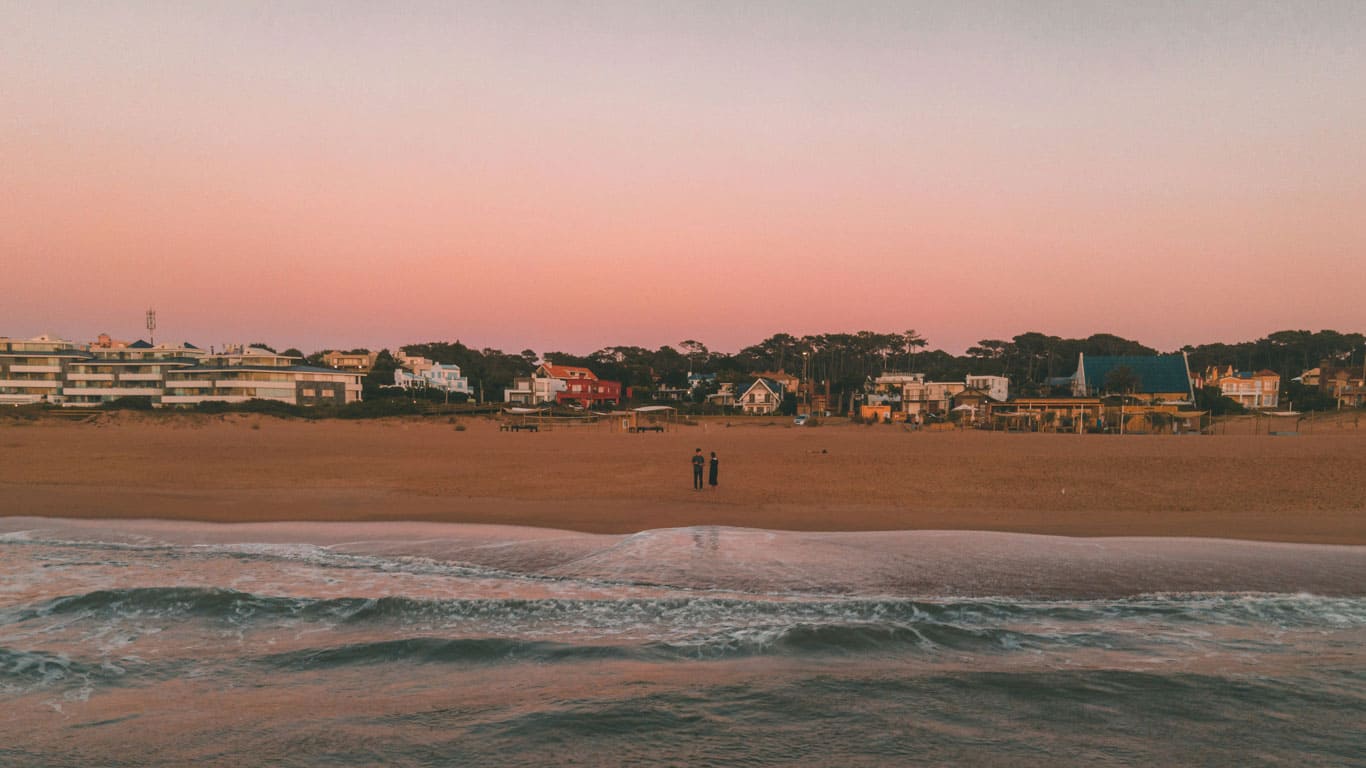 Gentle waves wash onto the sandy shores of La Barra beach in Punta del Este at dusk, with a couple standing near the water's edge and beachfront properties bathed in the soft pink hues of the setting sun."