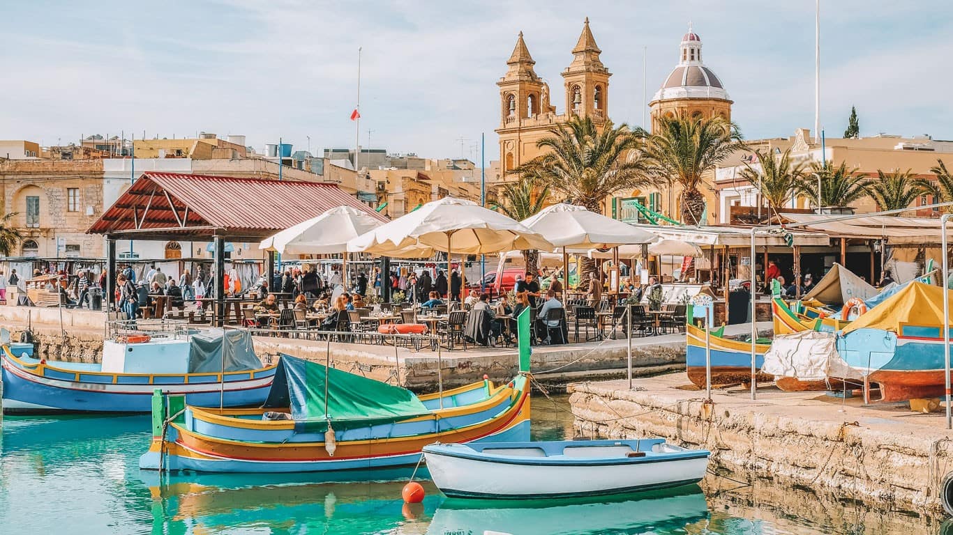 Vibrant harbor scene at Marsaxlokk in Malta, with the traditional brightly painted Maltese fishing boats, Luzzus, floating in the foreground, and a bustling market by the waterfront under parasols, framed by historical buildings and church spires.
