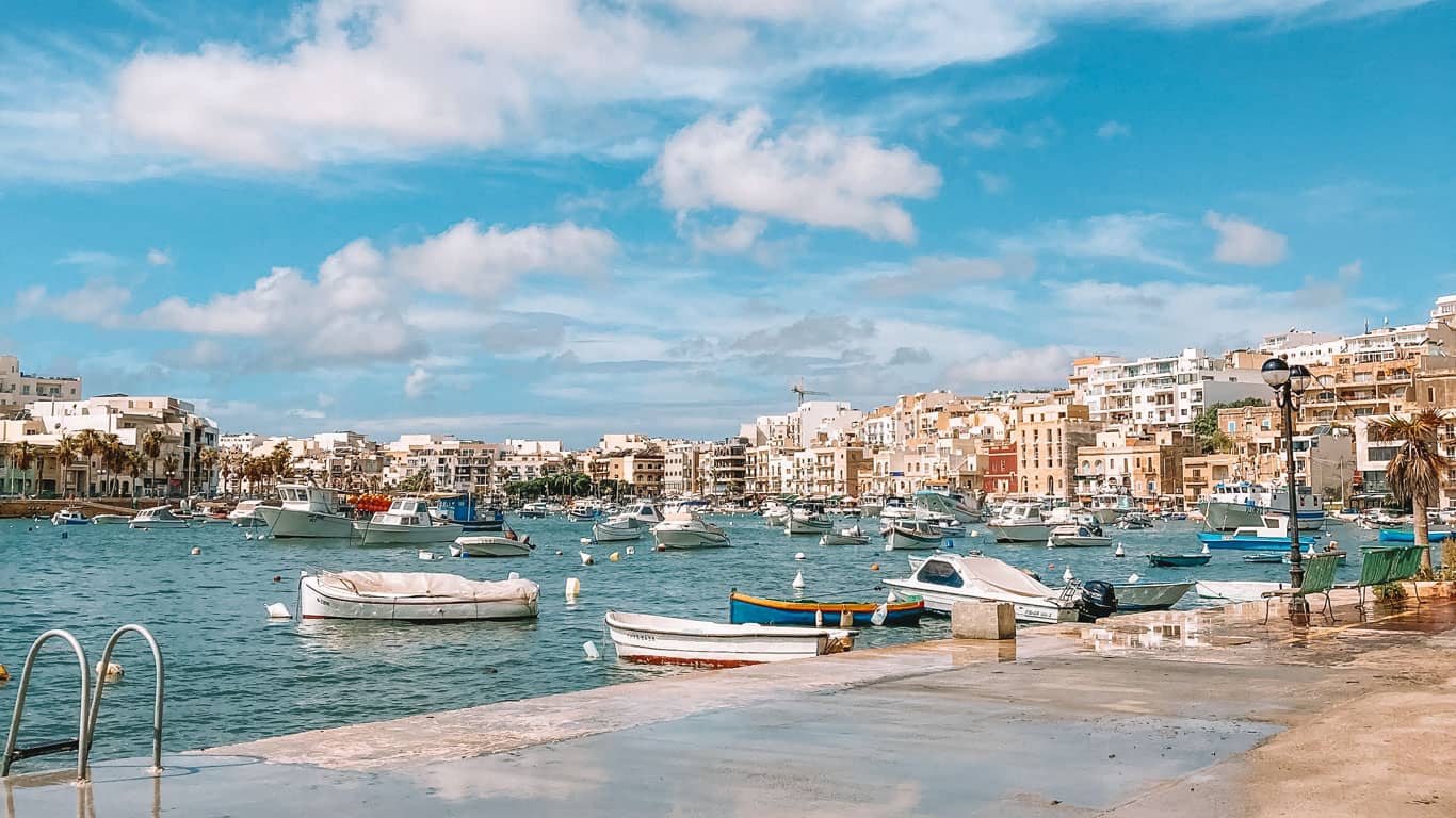 Bright and sunny day in Marsaskala, Malta, with a variety of boats moored in the bay's azure waters, complemented by the picturesque seafront lined with colorful Maltese architecture and leisurely palm trees, evoking a relaxed Mediterranean vibe.