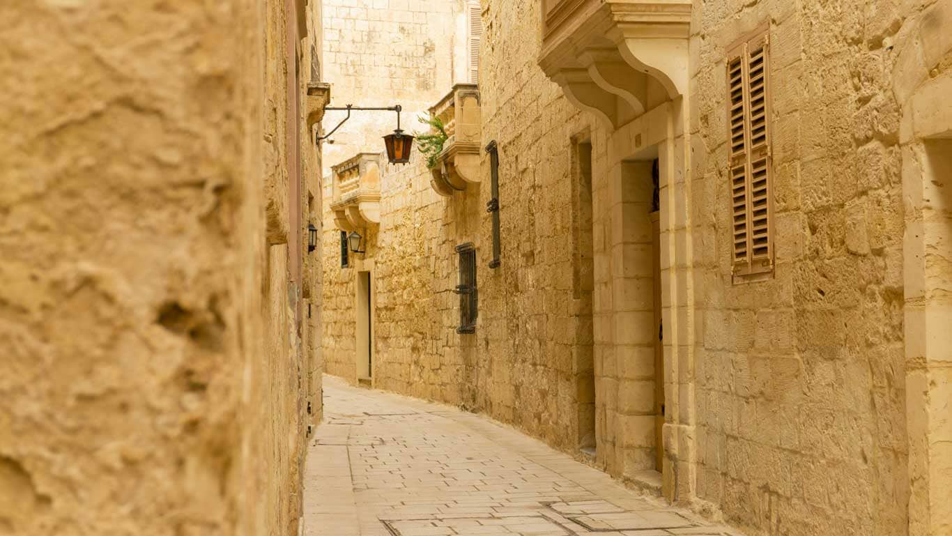A narrow alley in the ancient walled city of Mdina, Malta, lined with traditional limestone walls, adorned with vintage lanterns, and evoking a timeless atmosphere in the "Silent City."