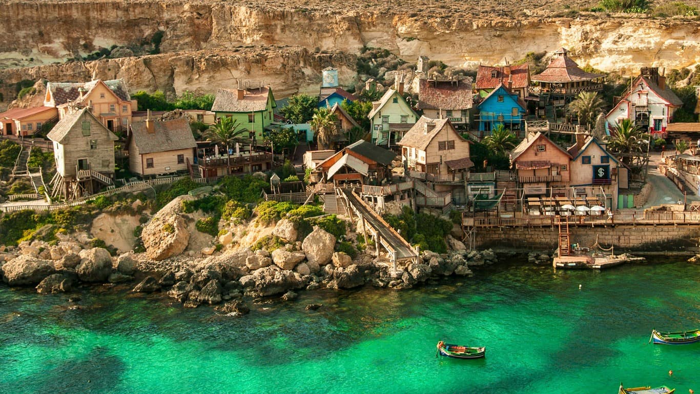Charming coastal village in Mellieha, Malta, with whimsical structures resembling a movie set, nestled against a rugged cliffside and overlooking clear emerald waters with traditional boats, capturing a fantasy-like Mediterranean atmosphere.