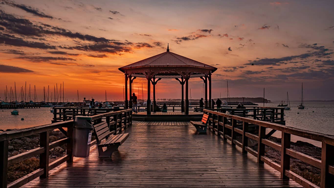 Twilight at the peninsula in Punta del Este, featuring a wooden pier leading to a gazebo with silhouetted figures enjoying the view, against a backdrop of moored sailboats under a vibrant orange sky