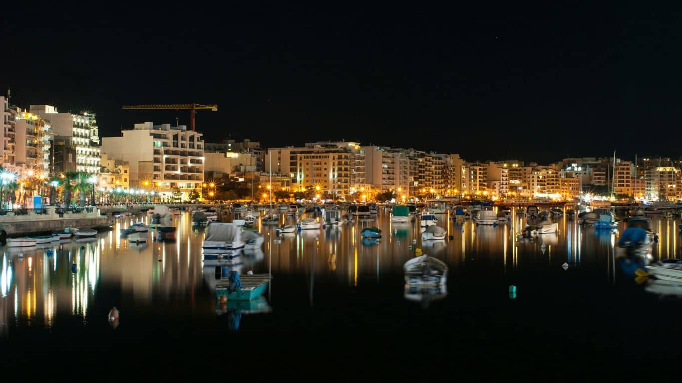 Nocturnal view of Sliema in Malta, with glowing city lights reflecting off the calm sea, dotted with moored boats creating a serene, yet vibrant waterfront scene.