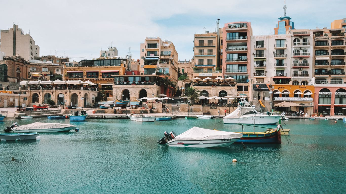 Colorful waterside scene at St. Julian's in Malta, featuring traditional Maltese boats bobbing in the aqua-marine waters, with a backdrop of quaint restaurants and multi-story buildings that capture the vibrant coastal life.