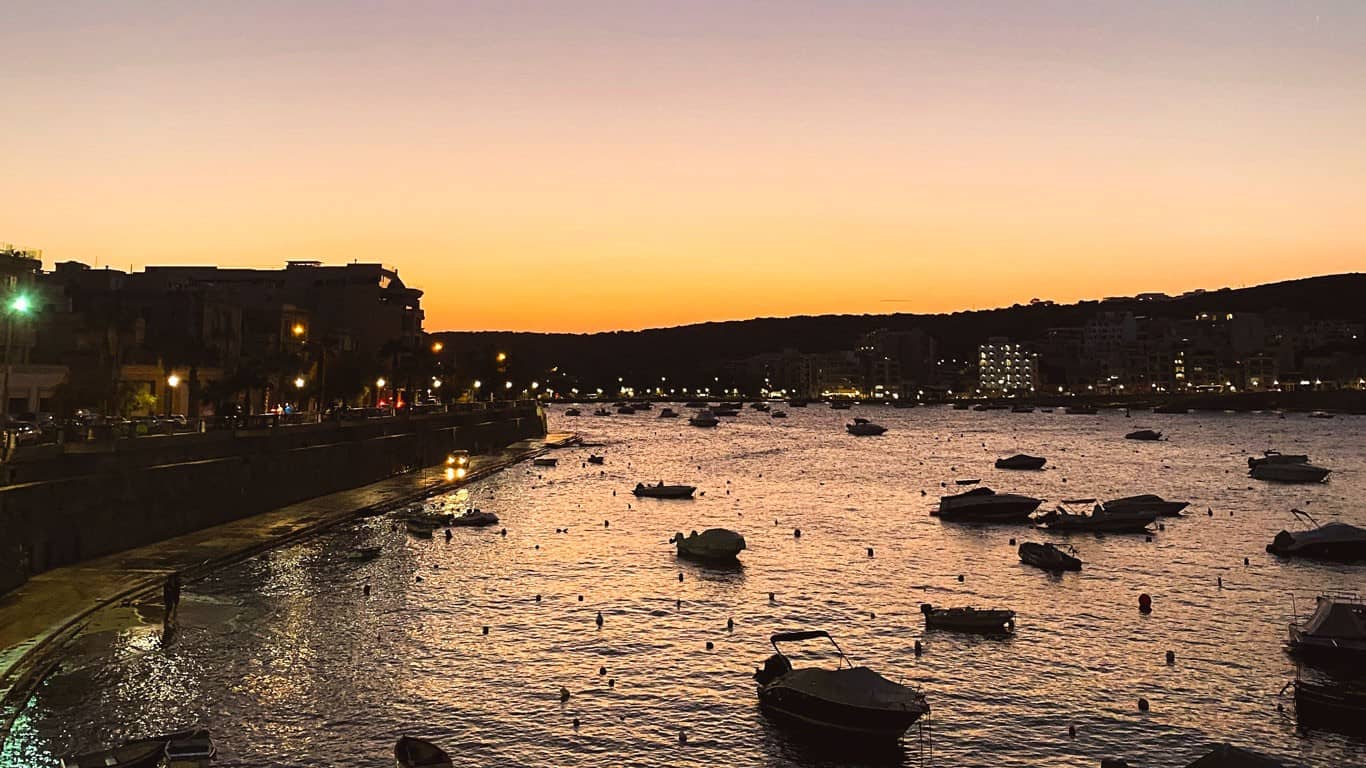 Twilight settles over St. Paul's Bay in Malta, with the last hues of the sunset casting a warm glow over the water, where boats gently sway, and the coastal promenade comes alive with evening lights.