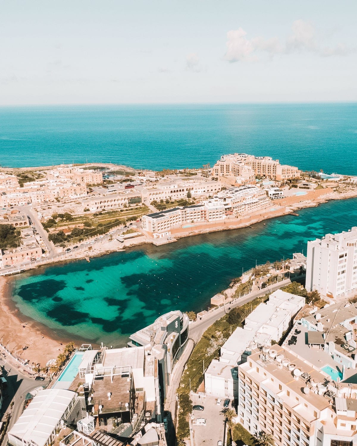 Aerial view of St. Julian's in Malta showcasing the clear turquoise waters of the bay, the surrounding Mediterranean architecture, and the urban landscape, with the caption highlighting it as the best place to stay.