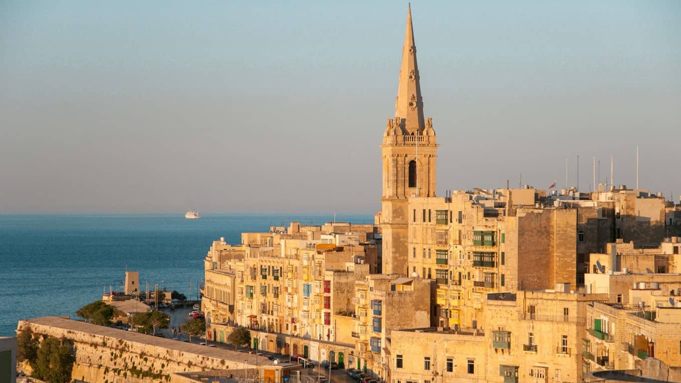 Sunlit view of Valletta in Malta, featuring the historic sandstone buildings with their traditional closed wooden balconies, and a striking church spire dominating the skyline, all set against the backdrop of the Mediterranean Sea.