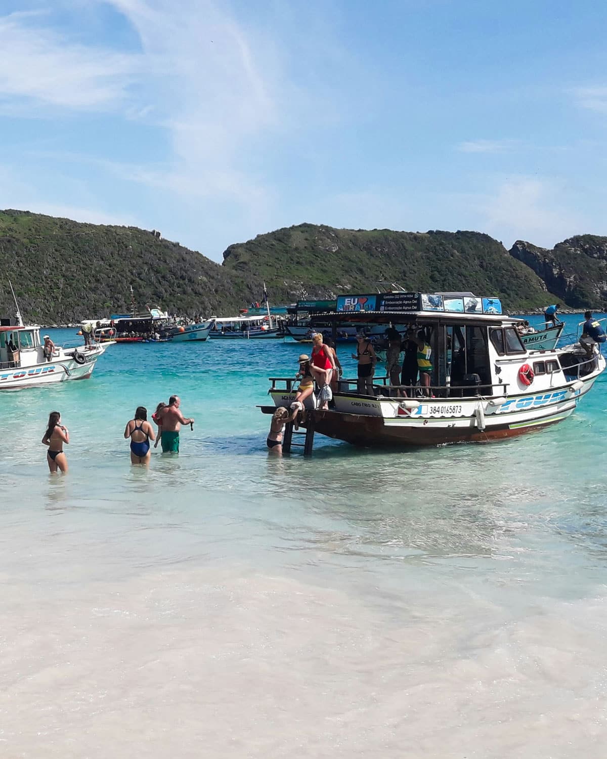Tourists enjoy the clear turquoise waters at Farol Beach in Arraial do Cabo, Brazil. Multiple boats are moored near the shore, with people disembarking into the shallow water. The scene captures a bustling day at one of the most popular beaches in Arraial do Cabo, surrounded by lush green hills under a bright blue sky.