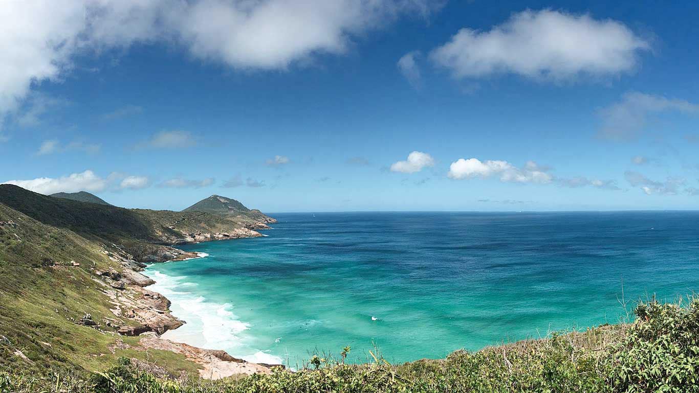 This image presents a panoramic view of Brava Beach in Arraial do Cabo, showcasing its rugged coastal beauty. The beach, nestled between rolling green hills, features pristine white sands and vibrant turquoise waters, contrasted by the rough, rocky coastline. The expansive blue sky above and the sparse cloud cover add to the serene and untouched appearance of this secluded beach.