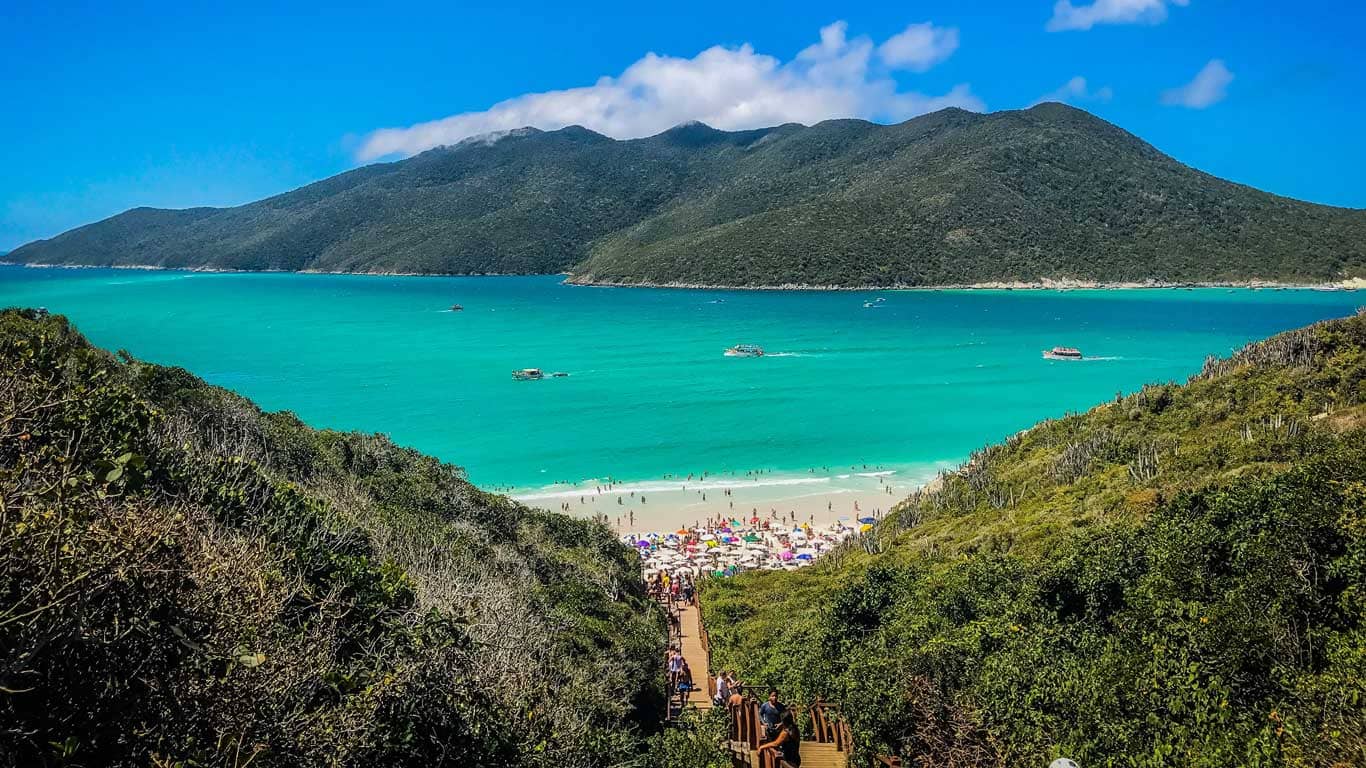 A breathtaking view from the top of a wooden staircase leading down to Prainhas do Pontal do Atalaia in Arraial do Cabo, Brazil. The beach is bustling with visitors enjoying the pristine white sands and vibrant turquoise waters, framed by lush green hills and dotted with boats. This panoramic scene highlights the natural beauty and popular appeal of one of Arraial do Cabo's most picturesque beaches.
