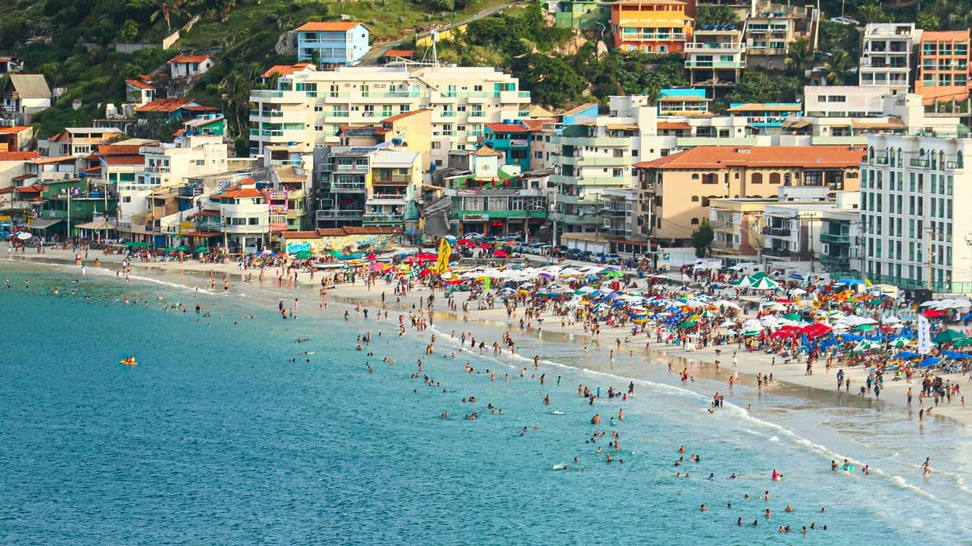 This vibrant image captures the bustling atmosphere of Prainha in Arraial do Cabo, Brazil. The beach is crowded with people, colorful umbrellas, and lively beach vendors, all set against a backdrop of tightly packed beachfront buildings that scale the surrounding hills. The crystal-clear blue waters provide a refreshing contrast to the dense urban landscape.